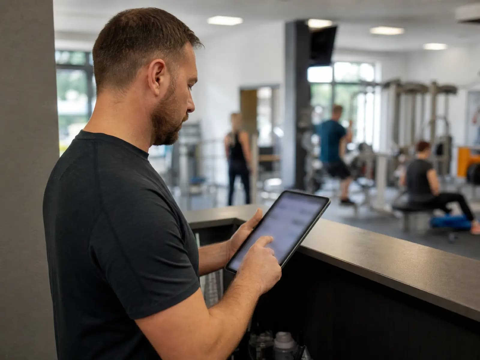 Trener przegląda listę klientów na tablecie przy recepcji siłowni